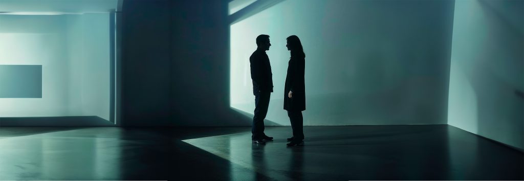 La foto muestra a dos figuras, un hombre y una mujer, de pie frente a frente en un amplio espacio interior moderno y minimalista. La iluminación tenue y las sombras alargadas crean un ambiente introspectivo. Ambas figuras están en silueta, lo que impide ver sus expresiones faciales o características distintivas, evocando un sentido de anonimato. El suelo reflejante y las paredes lisas sugieren una atmósfera fría y posiblemente aislante. La luz que entra a través de las aberturas geométricas de las paredes añade un elemento de diseño y podría simbolizar fuentes externas de influencia o información. La distancia física entre las dos personas, a pesar de su proximidad, puede interpretarse como una metáfora del "fin de lo social" en los medios sociales, donde las conexiones pueden ser superficiales y faltas de interacción humana verdadera.